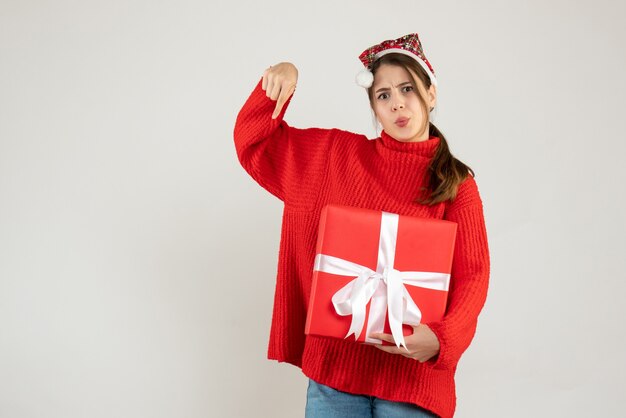 Front view confused girl with santa hat holding present finger pointing down standing