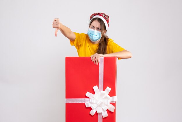 Front view confused girl with santa hat making thumb down sign standing behind big xmas gift