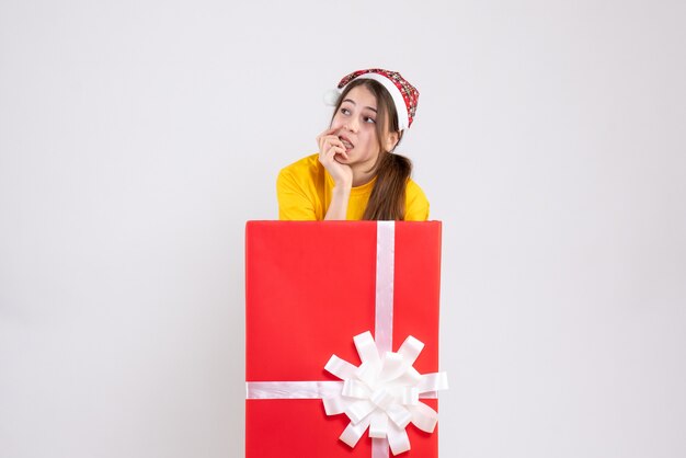 Front view confused girl with santa hat standing behind big xmas gift