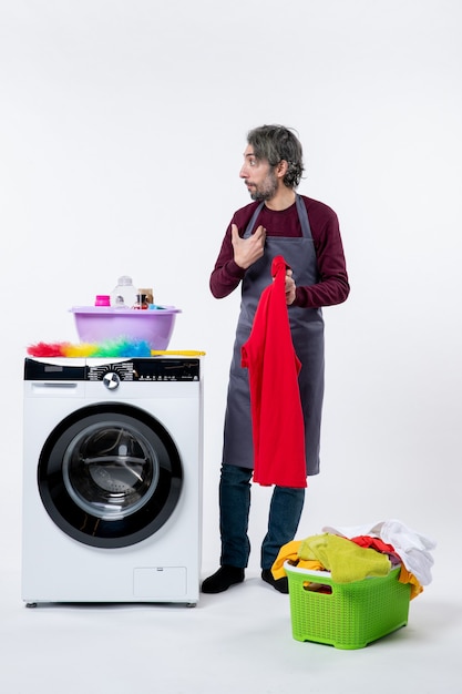 Front view confused housekeeper man holding red towel standing near washing machine on white background