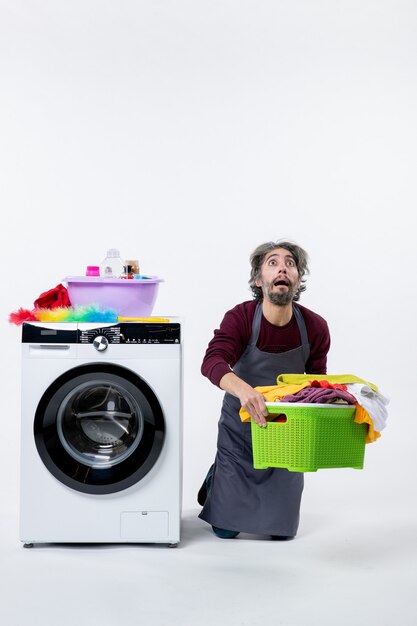 Front view confused housekeeper man kneeing near washer holding laundry basket on white background