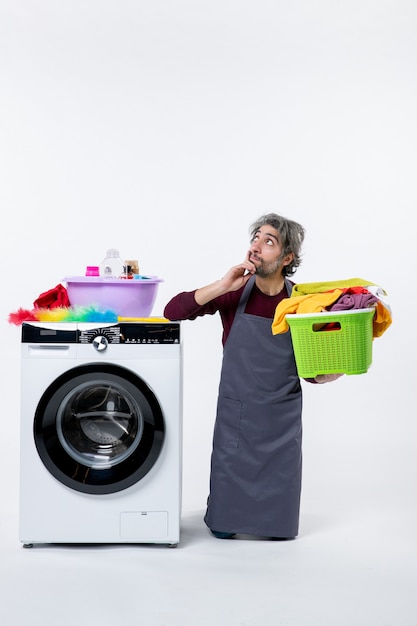 Free photo front view confused housekeeper man kneeing near washer holding up laundry basket on white background