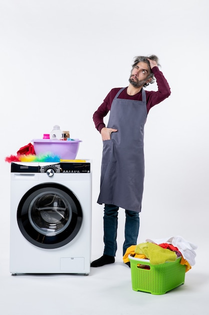 Front view confused housekeeper man putting hand in pocket standing near washing machine on white background