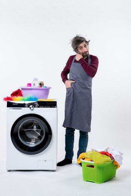 Front view confused housekeeper man putting hand in pocket standing near white washing machine on white background