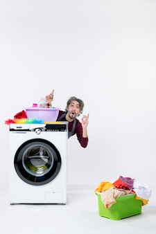 Front view confused male housekeeper in apron sitting behind washer laundry basket on white background