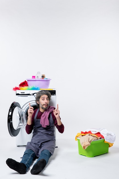 Front view confused male housekeeper reclining washer near laundry basket pointing at ceiling on white background