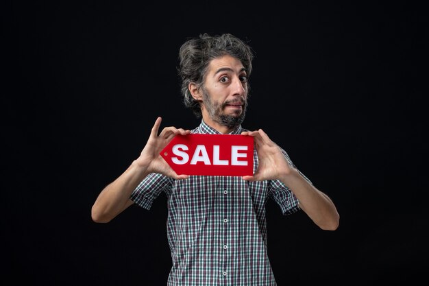 Front view confused man holding sale sign with bth hands on dark wall