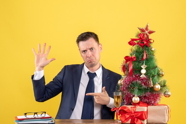 Front view of confused man opening his hand sitting at the table near xmas tree and presents on yellow