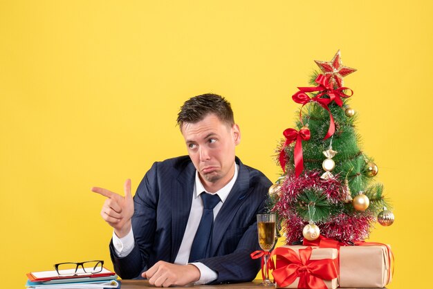 クリスマスツリーと黄色の贈り物の近くのテーブルに座っている何かを指している混乱した男の正面図