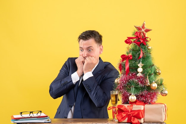 クリスマスツリーの近くのテーブルに座って、黄色で提示混乱した男の正面図