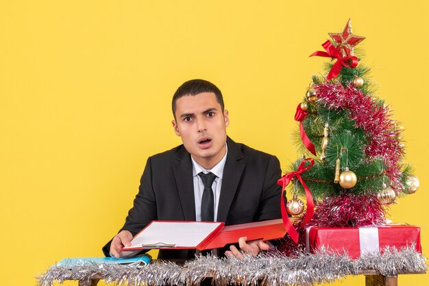 Front view confused man in suit sitting at the table holding document xmas tree and gifts