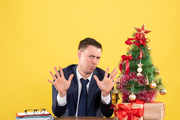 Front view of confused man with opened hands sitting at the table near xmas tree and presents on yellow