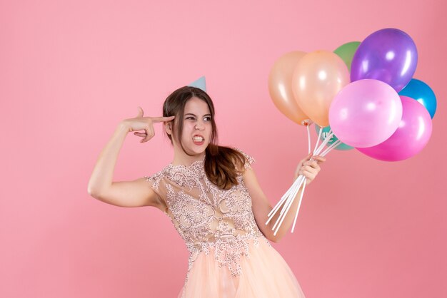 Front view confused party girl with party cap holding balloons keeping finger gun near her temple