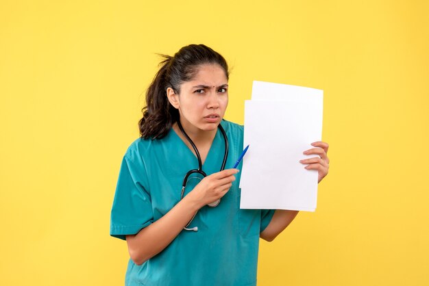 Front view confused pretty female doctor holding papers and pen on yellow background