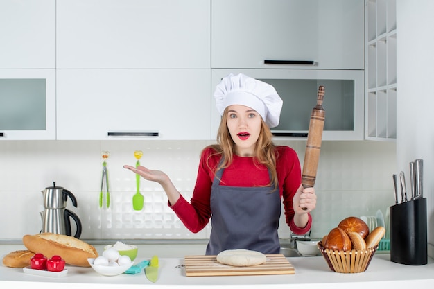 Front view confused woman in cook hat and apron holding rolling pin in the kitchen