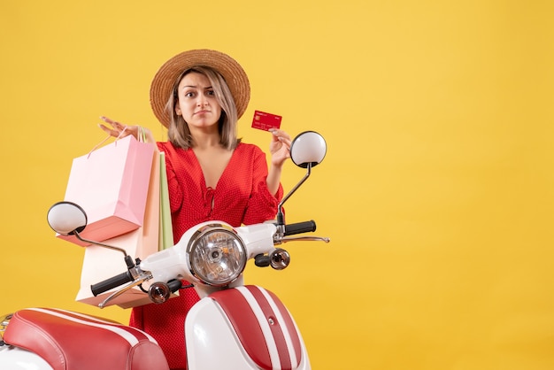 Front view of confused woman in red dress on moped holding shopping bags and card