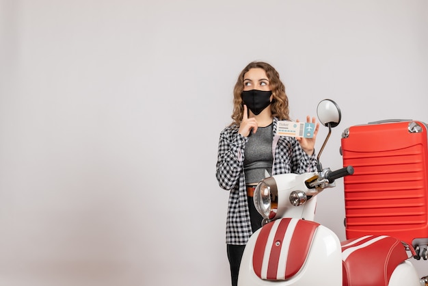 Front view of confused young girl with mask standing near moped with suitcase