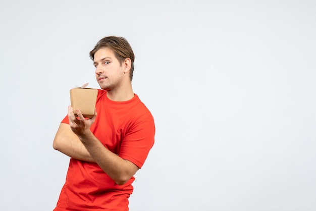 Вид спереди сбитого с толку молодого парня в красной блузке, держащего коробочку на белом фоне