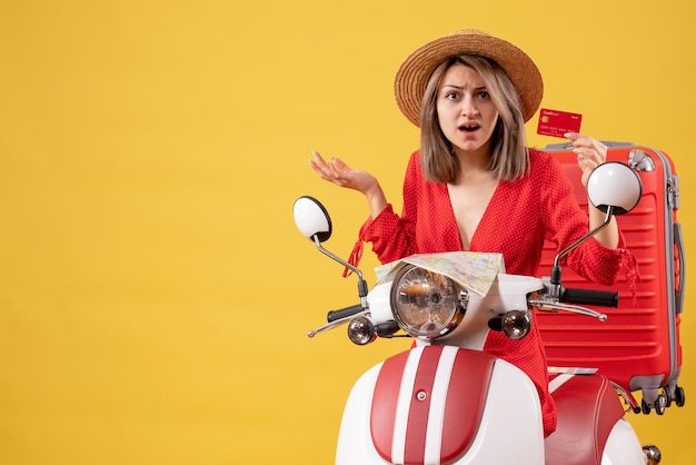 Front view of confused young lady in red dress holding card near moped