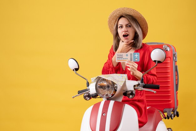 Front view of confused young lady in red dress holding ticket putting hand on her chin on moped