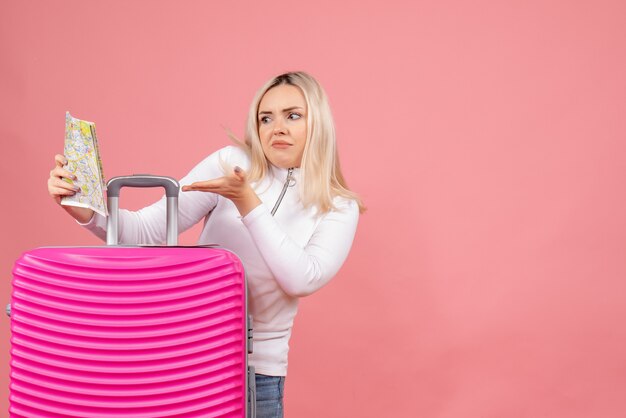 Front view confused young lady standing behind pink suitcase looking at map