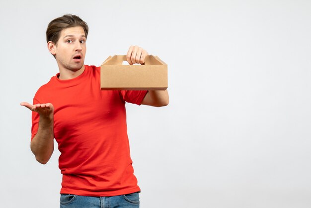 Front view of confused young man in red blouse holding box on white background