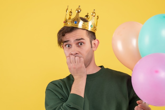 Front view confused young man with crown holding balloons on yellow 