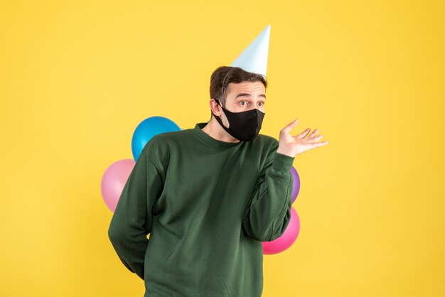 Front view confused young man with party cap and colorful balloons standing on yellow 