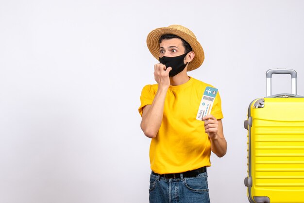 Front view confused young man with straw hat standing near yellow suitcase holding travel ticket