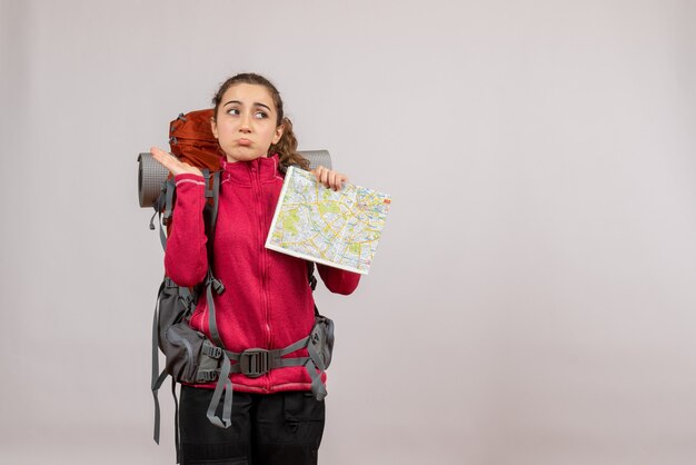 Front view of confused young traveller with big backpack holding up map on grey wall