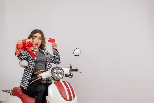 Front view of confused young woman on moped holding card and gift on grey wall