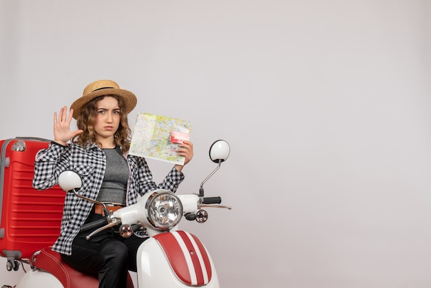 Front view of confused young woman on moped holding card and map on grey wall