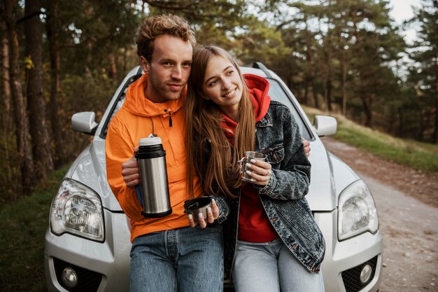 屋外で車のボンネットに座って温かい飲み物を楽しんでいるカップルの正面図