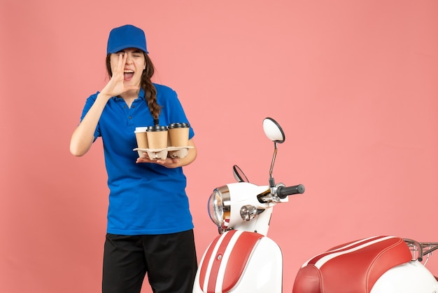 パステル ピーチ色の背景に誰かを呼び出すコーヒーを保持しているオートバイの隣に立っている宅配便の女の子の正面図