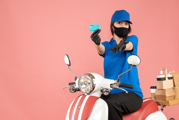 Front view of courier girl wearing medical mask and gloves sitting on scooter holding bank card delivering orders on pastel peach background