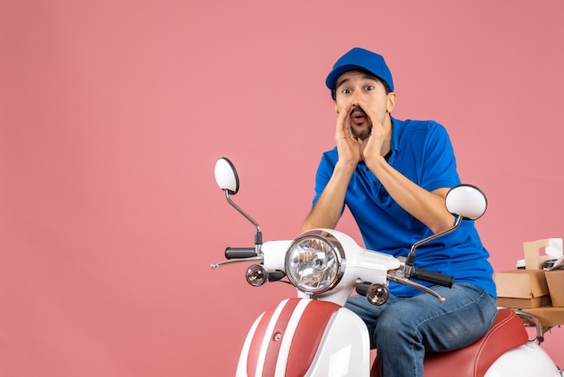 Front view of courier guy wearing hat sitting on scooter calling someone on pastel peach background