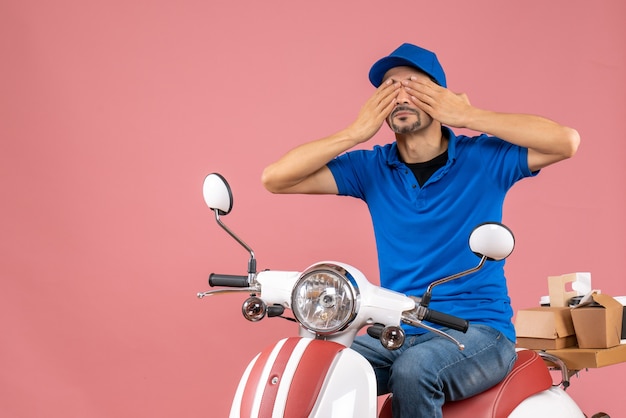 Front view of courier guy wearing hat sitting on scooter closing his eyes on pastel peach background