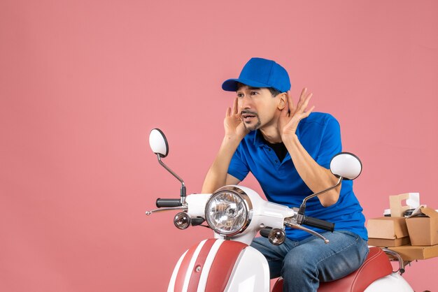 Front view of courier guy wearing hat sitting on scooter listening to the last gossiping on pastel peach background