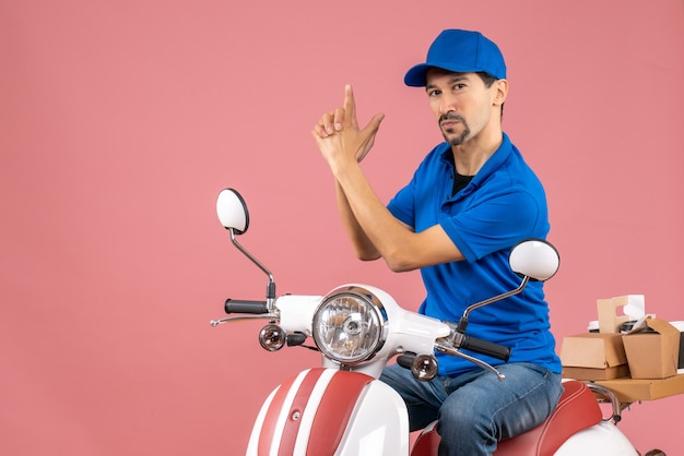 Front view of courier guy wearing hat sitting on scooter and making gun gesture on pastel peach background