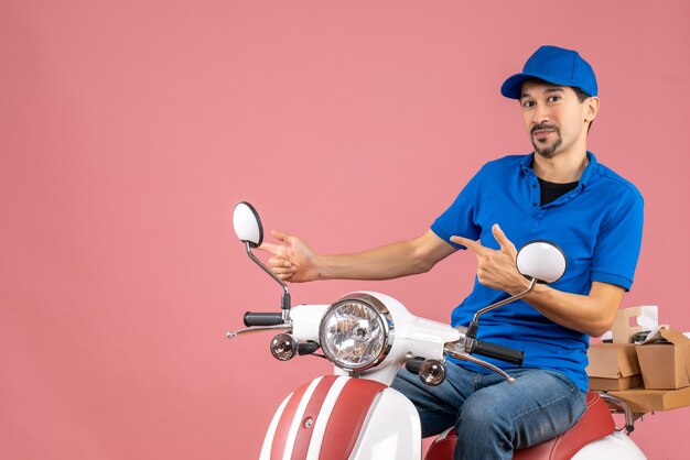 Front view of courier guy wearing hat sitting on scooter pointing something on the right side on pastel peach background