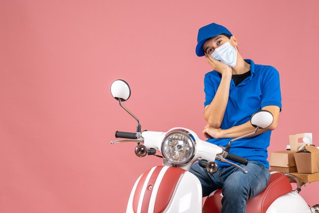 Front view of courier man in medical mask wearing hat sitting on scooter and looking at something with hopeful facial expression on pastel peach background