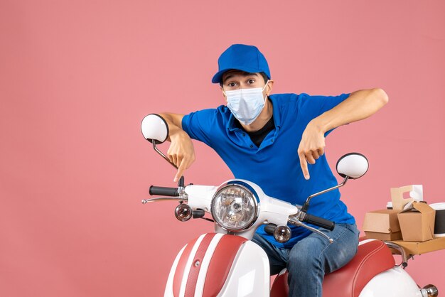 Front view of courier man in medical mask wearing hat sitting on scooter and pointing down on pastel peach background