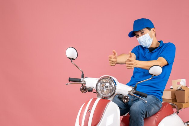 Front view of courier man in medical mask wearing hat sitting on scooter and pointing forward on pastel peach background