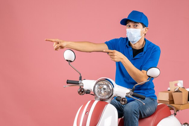 Front view of courier man in medical mask wearing hat sitting on scooter and pointing something on the right side on pastel peach background