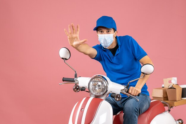 Vista frontale dell'uomo corriere in maschera medica che indossa un cappello seduto su uno scooter che mostra cinque su sfondo color pesca pastello