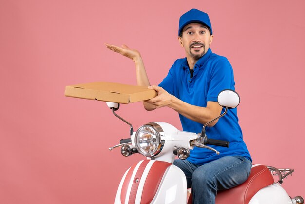 Front view of courier man wearing hat sitting on scooter holding order and making exact something on pastel peach background