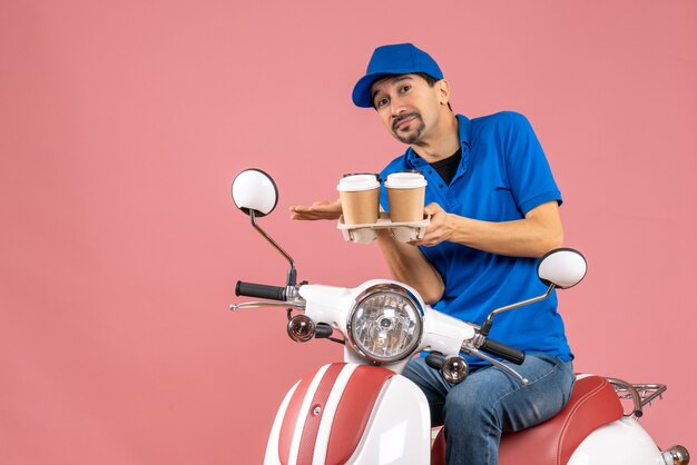 Front view of courier man wearing hat sitting on scooter making exact something on pastel peach background