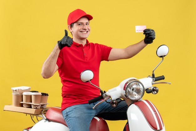 Вид спереди курьера в красной блузке и шляпных перчатках в медицинской маске, доставляющего заказ, сидя на скутере с банковской картой и делая жест `` ОК ''