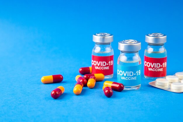 青い波の背景の左側にあるCOVID-ワクチンアンプルとカプセルが詰められた錠剤の正面図