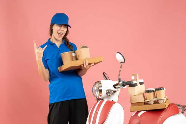 Front view of crazy emotional courier girl standing next to motorcycle holding coffee and small cakes on pastel peach color background